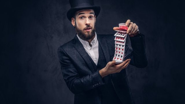 un-mago-barbudo-con-traje-negro-y-sombrero-de-copa-mostrando-trucos-con-cartas-en-un-fondo-oscuro